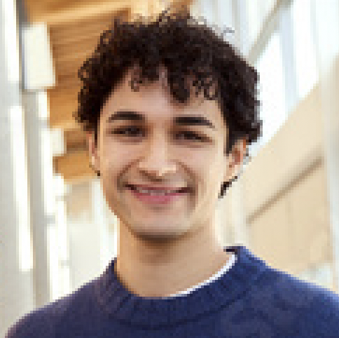 A young person with short, curly dark hair smiles while wearing a blue sweater, standing indoors in a bright hallway with large windows.