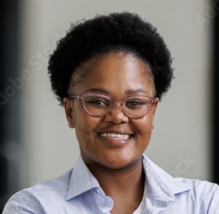 A person with short curly hair, wearing glasses and a light plaid shirt, smiling while standing indoors against a blurred background.