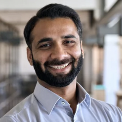 A man with a beard and mustache smiles warmly, wearing a light blue collared shirt. The background is softly blurred, suggesting an indoor office or library setting.