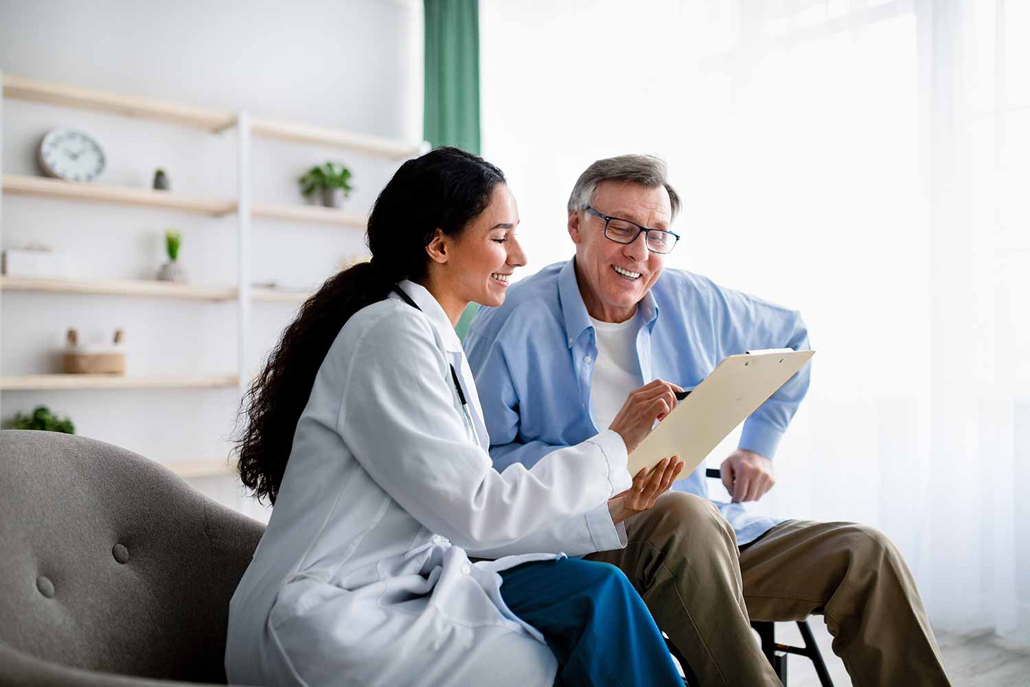 A smiling doctor shows a clipboard to an older man who is also smiling. They are sitting together in a bright, comfortable room with shelves and a clock in the background.