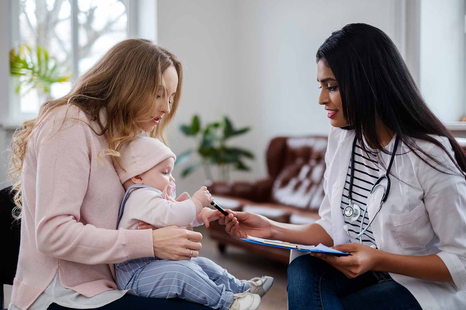 A female doctor hands a pen to a baby sitting on a womans lap during a consultation at home. The woman watches the baby, and a clipboard is visible in the doctors hand.