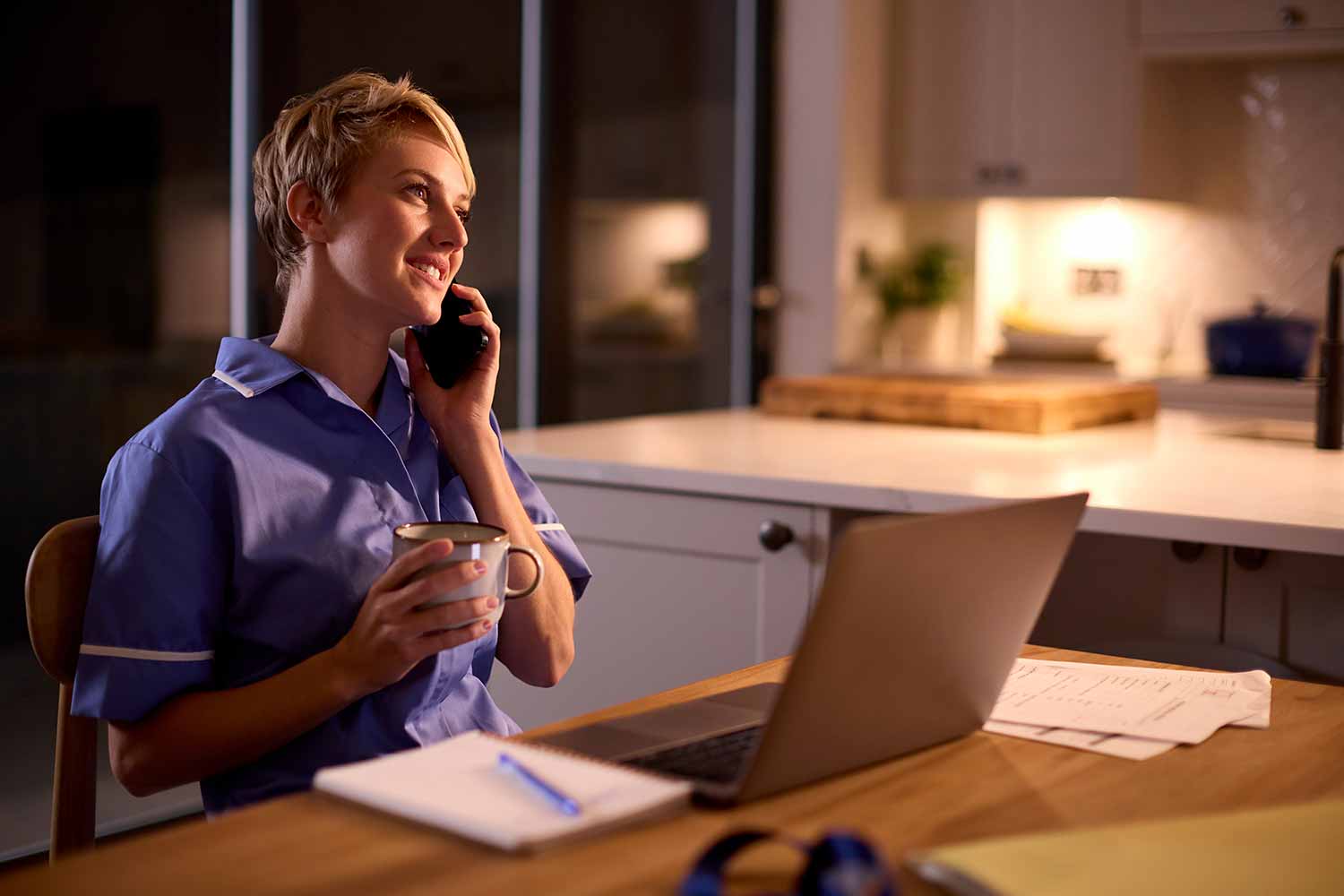 A woman in scrubs sits at a kitchen table, smiling while talking on the phone. She holds a mug and has a laptop, notebook, and papers in front of her, suggesting she is working or studying from home in the evening.