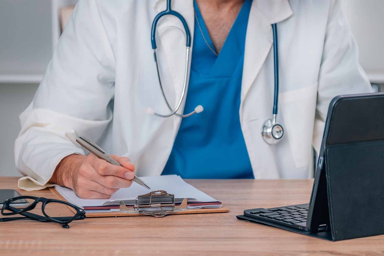 A doctor in a white coat writes on a clipboard at a desk with a laptop and eyeglasses nearby.