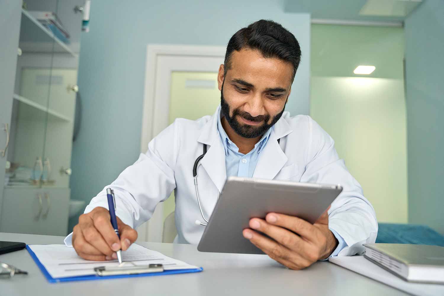 Male doctor in a white coat sits at a desk, smiling while writing on a clipboard and holding a tablet. Medical equipment and shelves are visible in the background.