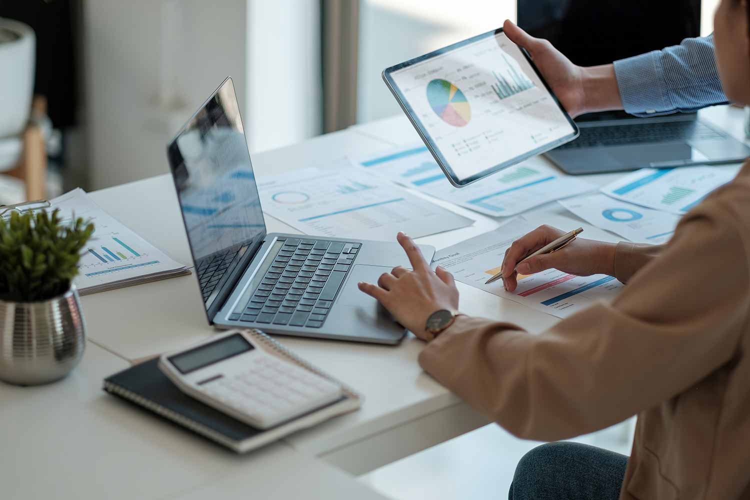 Two people work at a desk with laptops, charts, papers, and a calculator, analyzing data; one person holds a tablet displaying colorful graphs and charts.