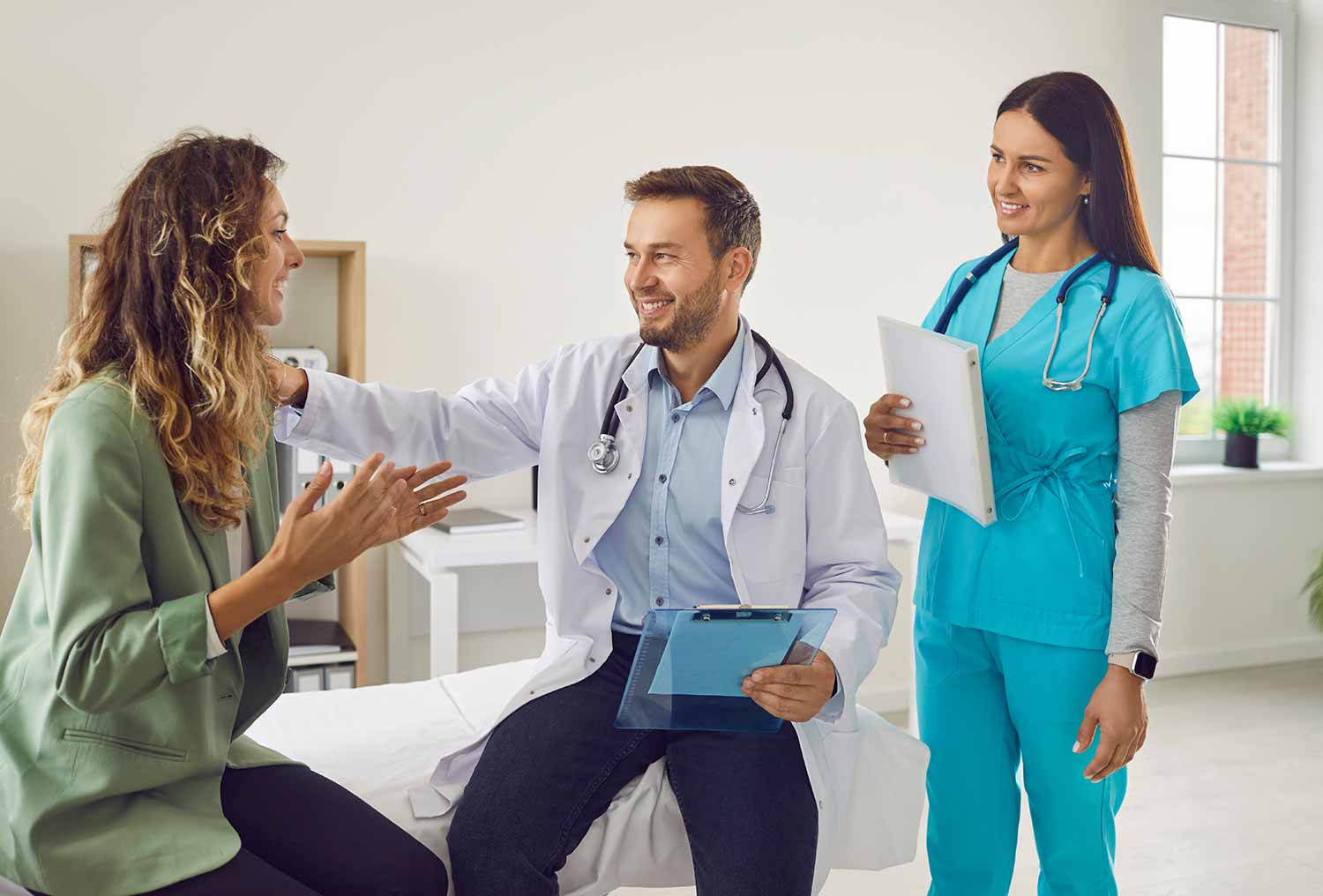 A doctor and nurse smiling while talking with a patient in a medical office. The patient is sitting on an exam table, gesturing happily. The nurse holds a clipboard and all appear engaged in a positive conversation.