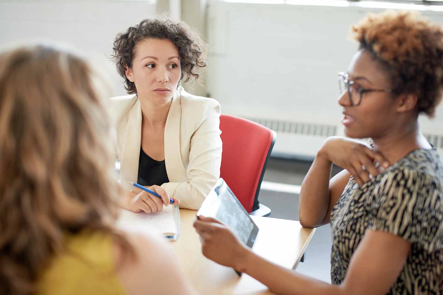 Three women sit at a table in discussion. One woman holds a tablet while talking; another listens attentively with a pen in hand. The third womans back faces the camera.